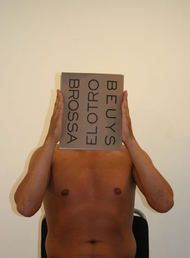 Peniche, sentado, se tapa el rostro con el libro «El otro», con los nombres de Brossa y Beuys en la portada